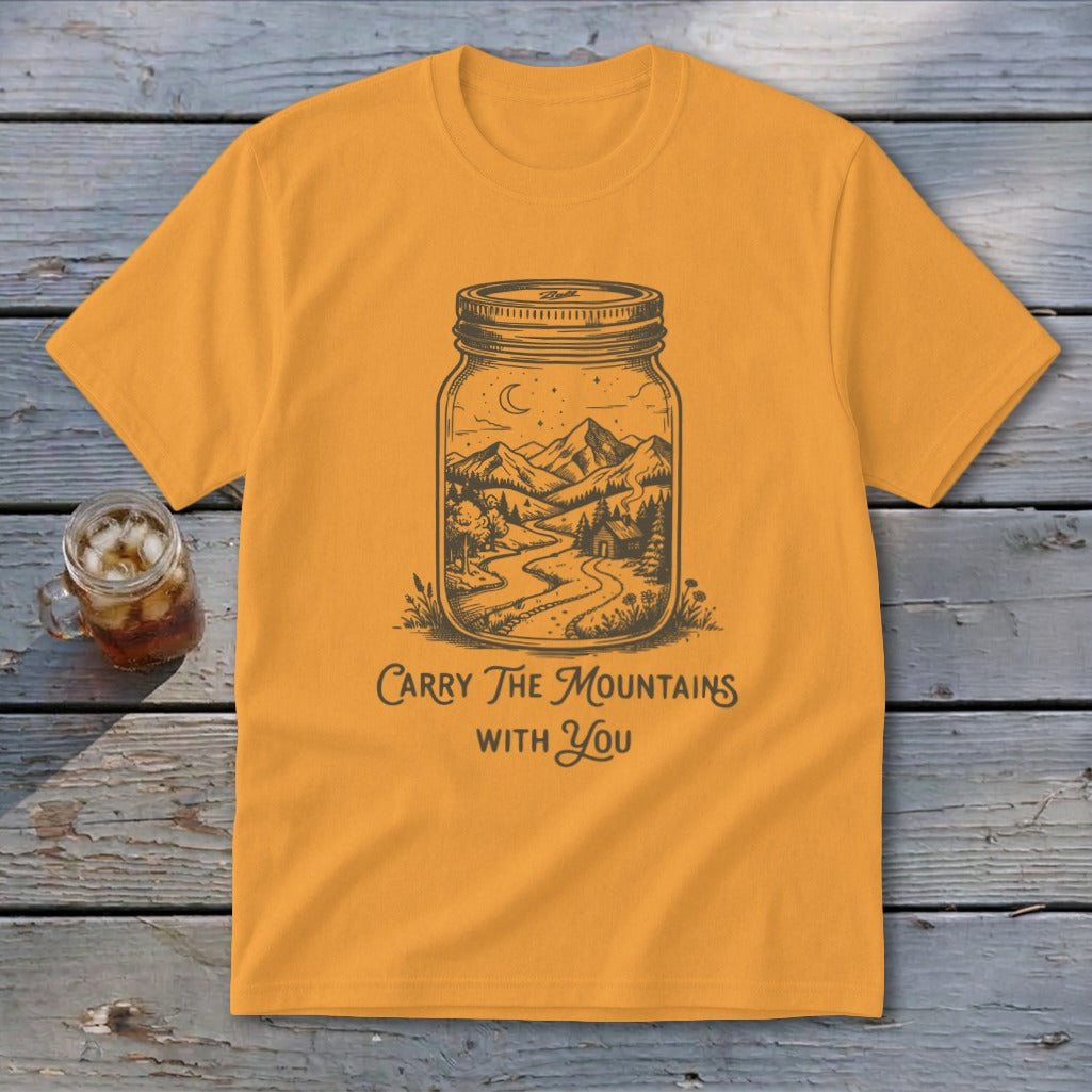 A mustard yellow t-shirt features a black illustration of a mountain landscape inside a mason jar, with the text "Carry The Mountains With You."