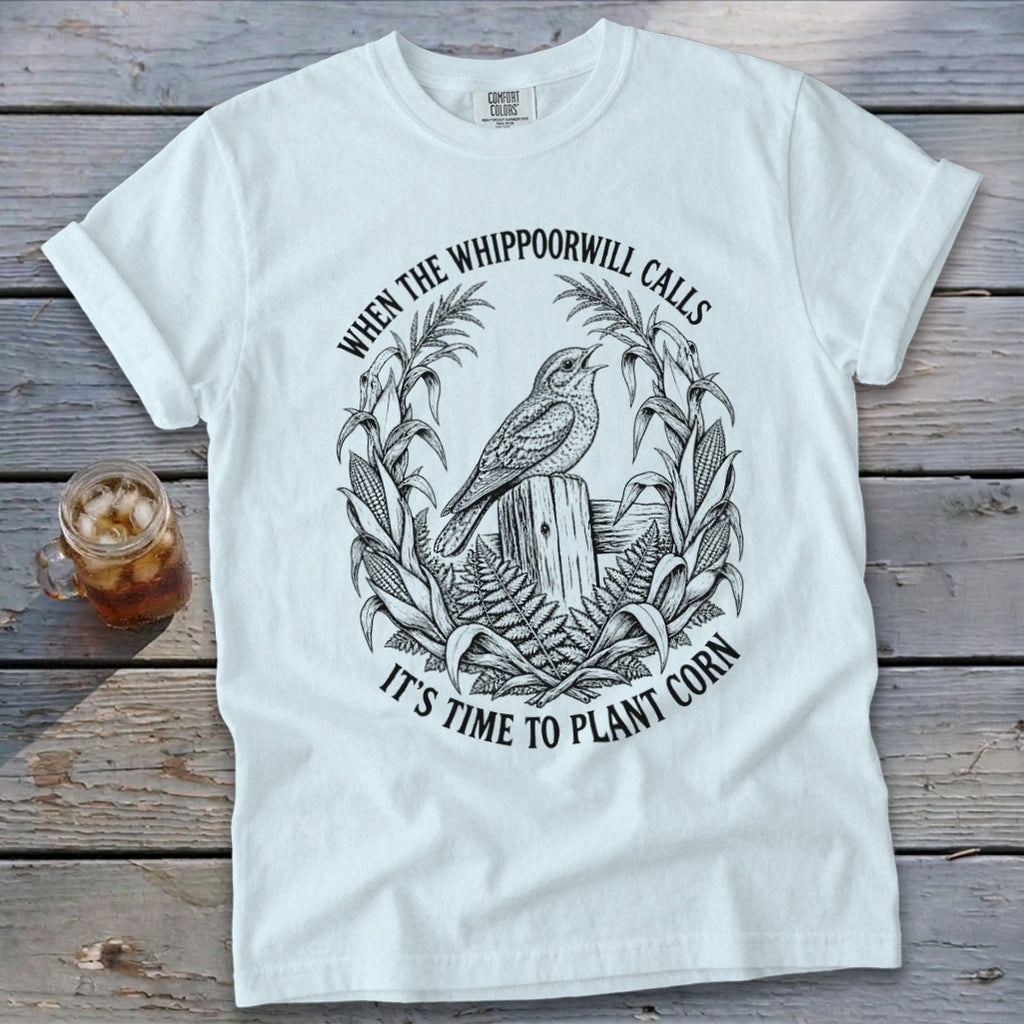 A white t-shirt featuring a black graphic of a bird, corn stalks, and text reading "When the whippoorwill calls it's time to plant corn".
