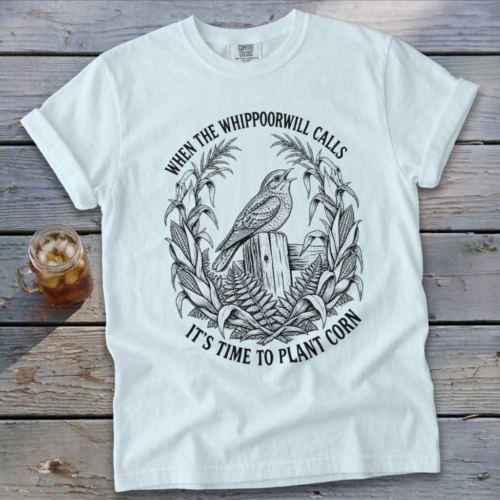 A white t-shirt featuring a black graphic of a bird, corn stalks, and text reading "When the whippoorwill calls it's time to plant corn".