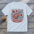 A white t-shirt featuring a colorful graphic of an acoustic guitar, sunflowers, and the text MOUNTAIN MELODIES.