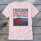 A pink crewneck t-shirt featuring a mountain graphic and the text FREEDOM RINGS LOUDEST IN THESE HILLS.
