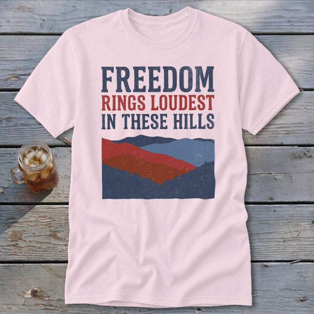 A pink crewneck t-shirt featuring a mountain graphic and the text FREEDOM RINGS LOUDEST IN THESE HILLS.