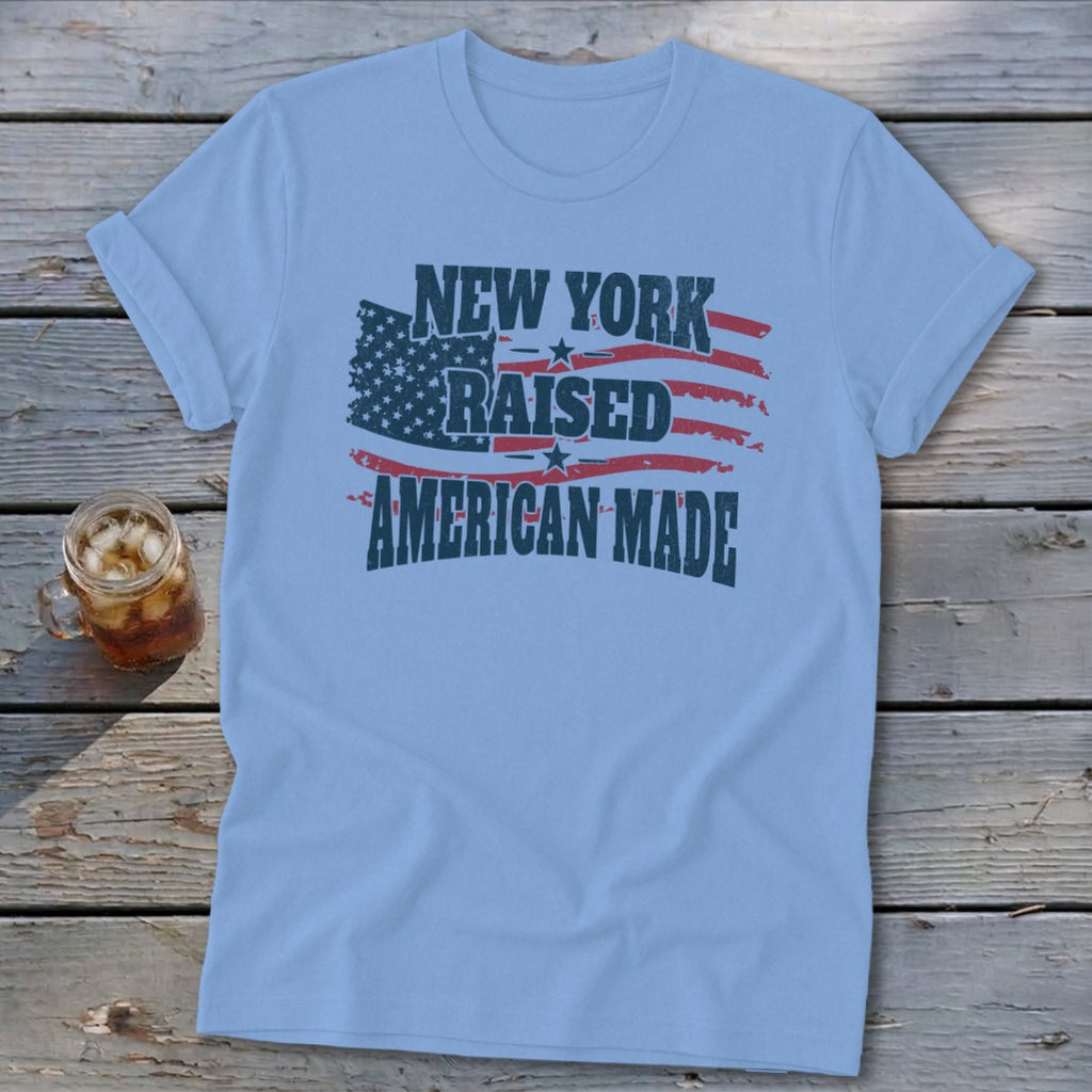 Light blue crewneck t-shirt with rolled sleeves laid flat on a rustic wooden surface, featuring text that reads New York Raised American Made over an American flag graphic.