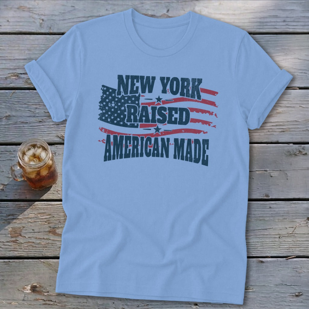 Light blue crewneck t-shirt with rolled sleeves laid flat on a rustic wooden surface, featuring text that reads New York Raised American Made over an American flag graphic.