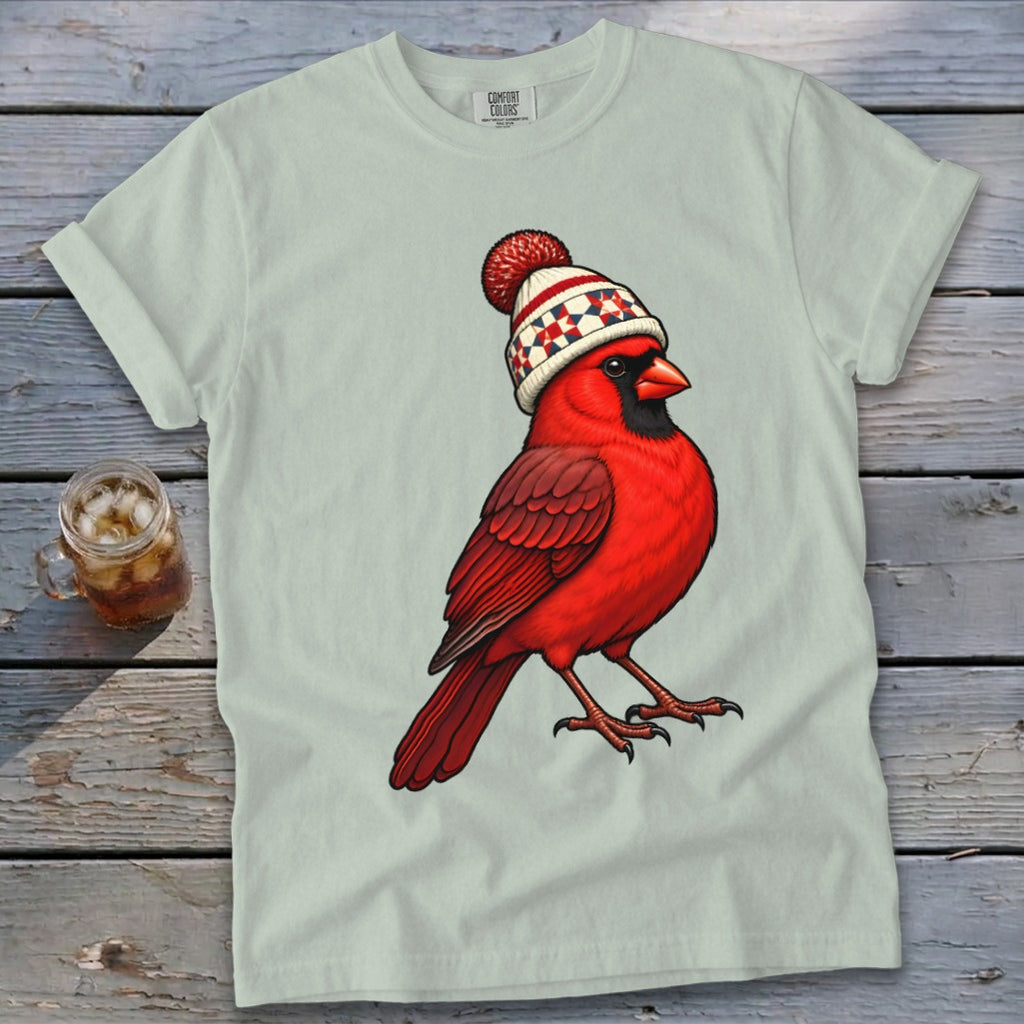 Light gray t-shirt featuring a cartoon red cardinal wearing a patterned winter hat, lying on a wooden surface.