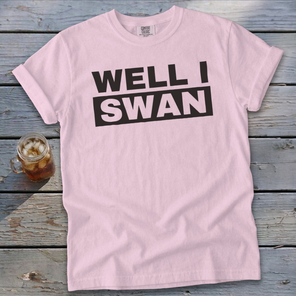 A light pink t-shirt with black text reading WELL I SWAN lies flat on a weathered wooden surface next to a glass of iced tea.
