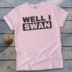 A light pink t-shirt with black text reading WELL I SWAN lies flat on a weathered wooden surface next to a glass of iced tea.