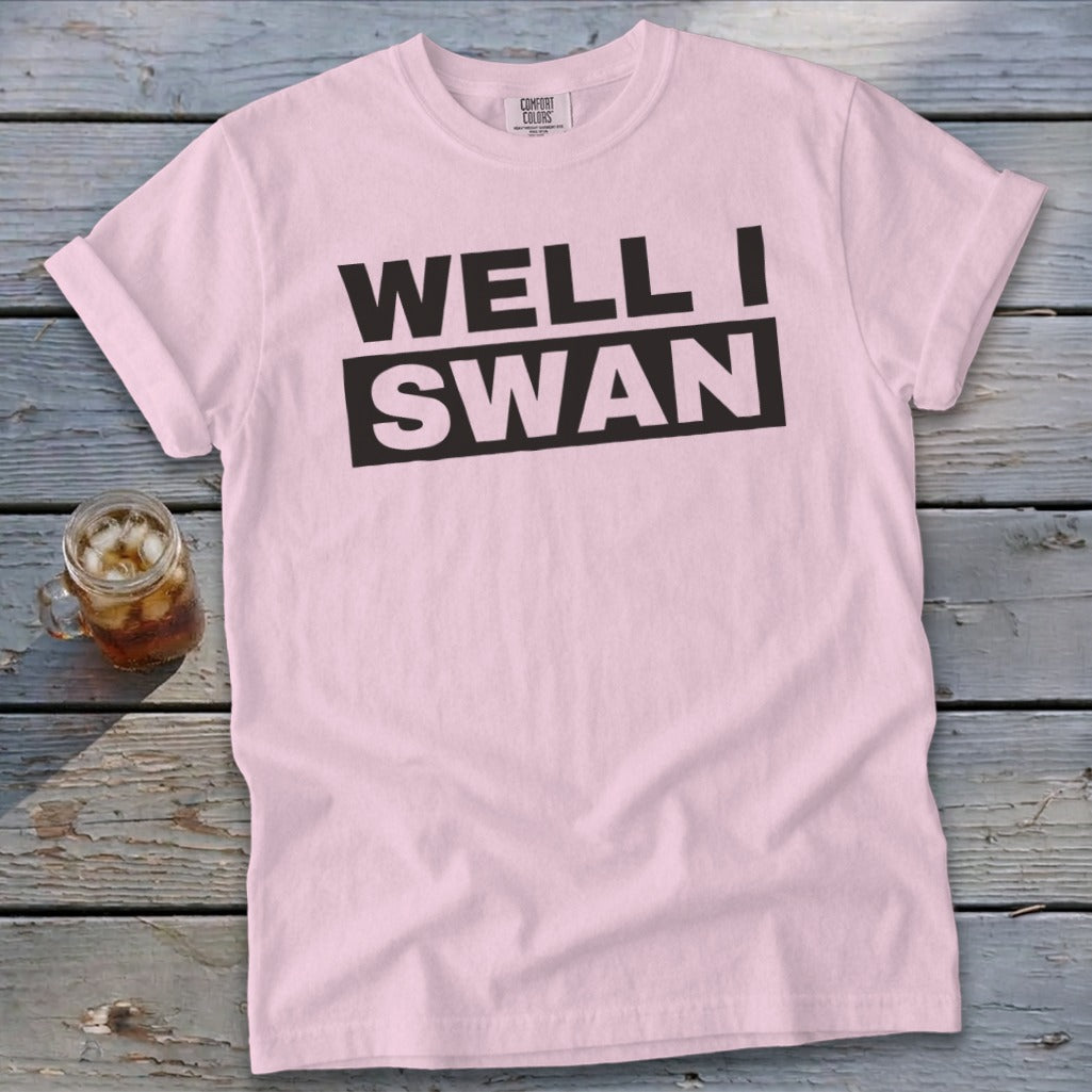 A light pink t-shirt with black text reading WELL I SWAN lies flat on a weathered wooden surface next to a glass of iced tea.