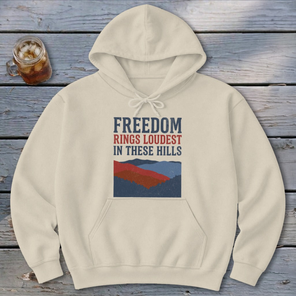 A cream-colored hoodie features a graphic of overlapping hills and text reading FREEDOM RINGS LOUDEST IN THESE HILLS.