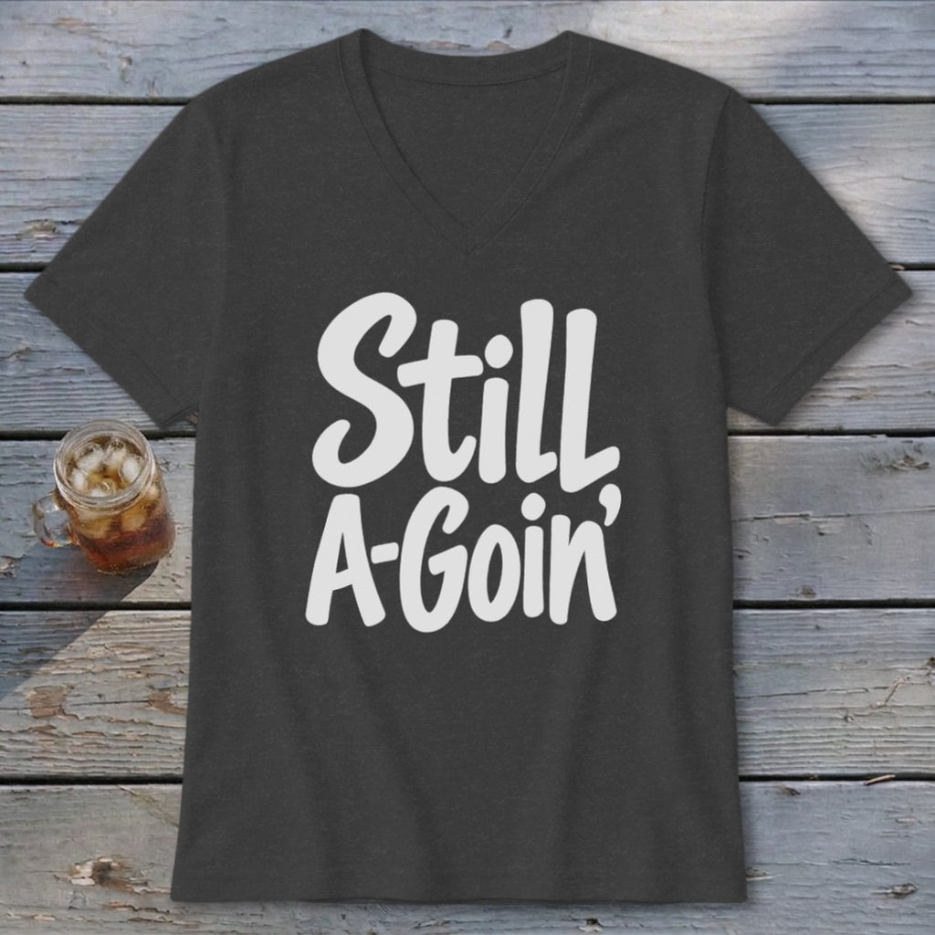 A charcoal V-neck t-shirt with white text reading Still A-Goin' on a wooden background.