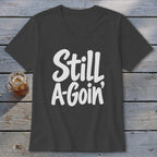 A charcoal V-neck t-shirt with white text reading Still A-Goin' on a wooden background.