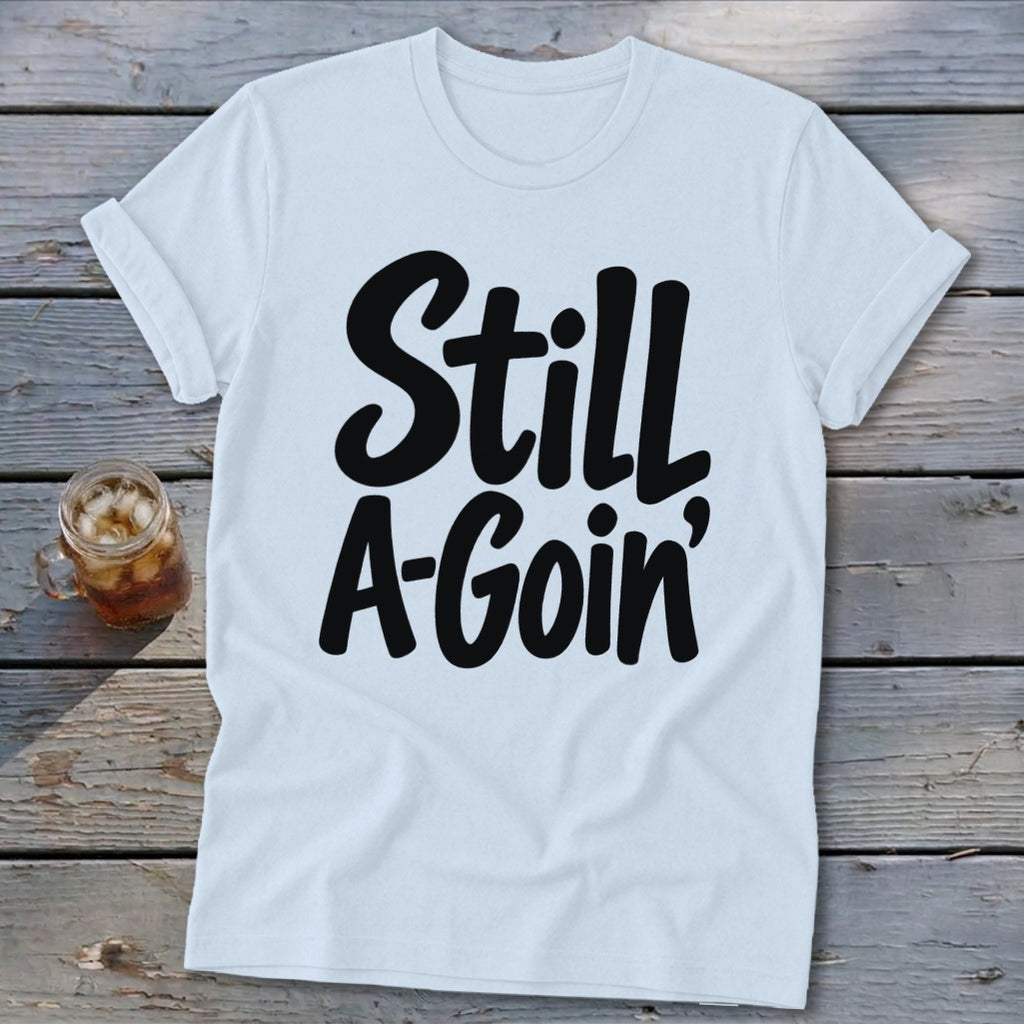 A white short-sleeved t-shirt with rolled cuffs rests on a wooden surface, featuring the black text "Still A-Goin'".