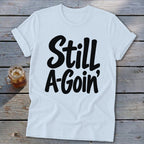 A white short-sleeved t-shirt with rolled cuffs rests on a wooden surface, featuring the black text "Still A-Goin'".