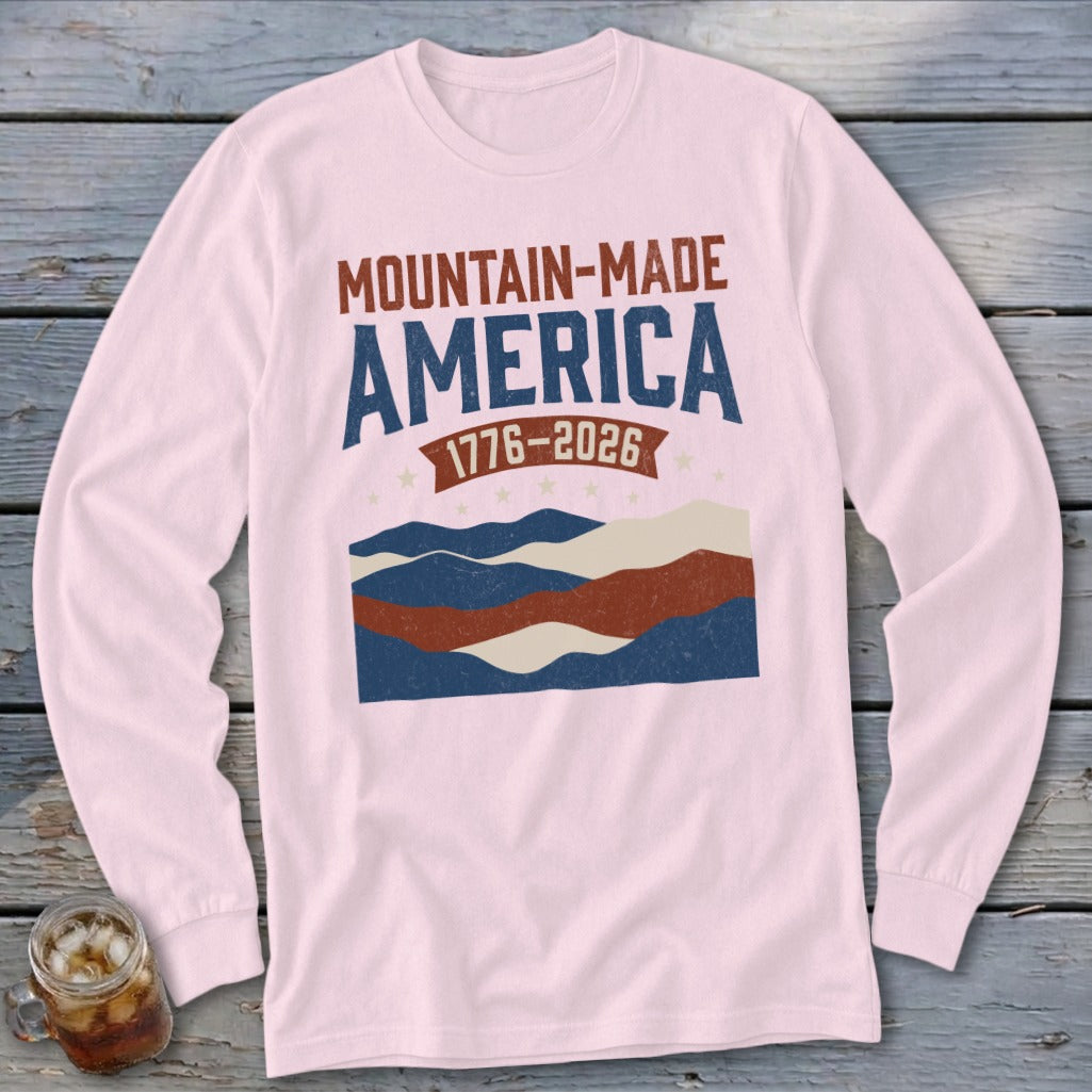 A light pink long-sleeve shirt featuring a graphic with the text Mountain-Made America 1776-2026 over abstract mountains.
