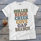 White crewneck t-shirt with rolled sleeves laid on wood, featuring stacked multi-colored text reading HOLLER RIDGE CREEK COVE GAP BRANCH.