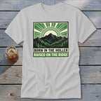 Light gray t-shirt with a green graphic of mountains, sun rays, and text that reads Born in the Holler, Raised on the Ridge.