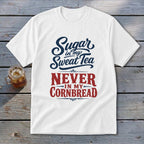 A white short-sleeved t-shirt with blue and red text that reads Sugar in my Sweat Tea NEVER IN MY CORNBREAD rests on a wooden surface beside a glass of iced tea.