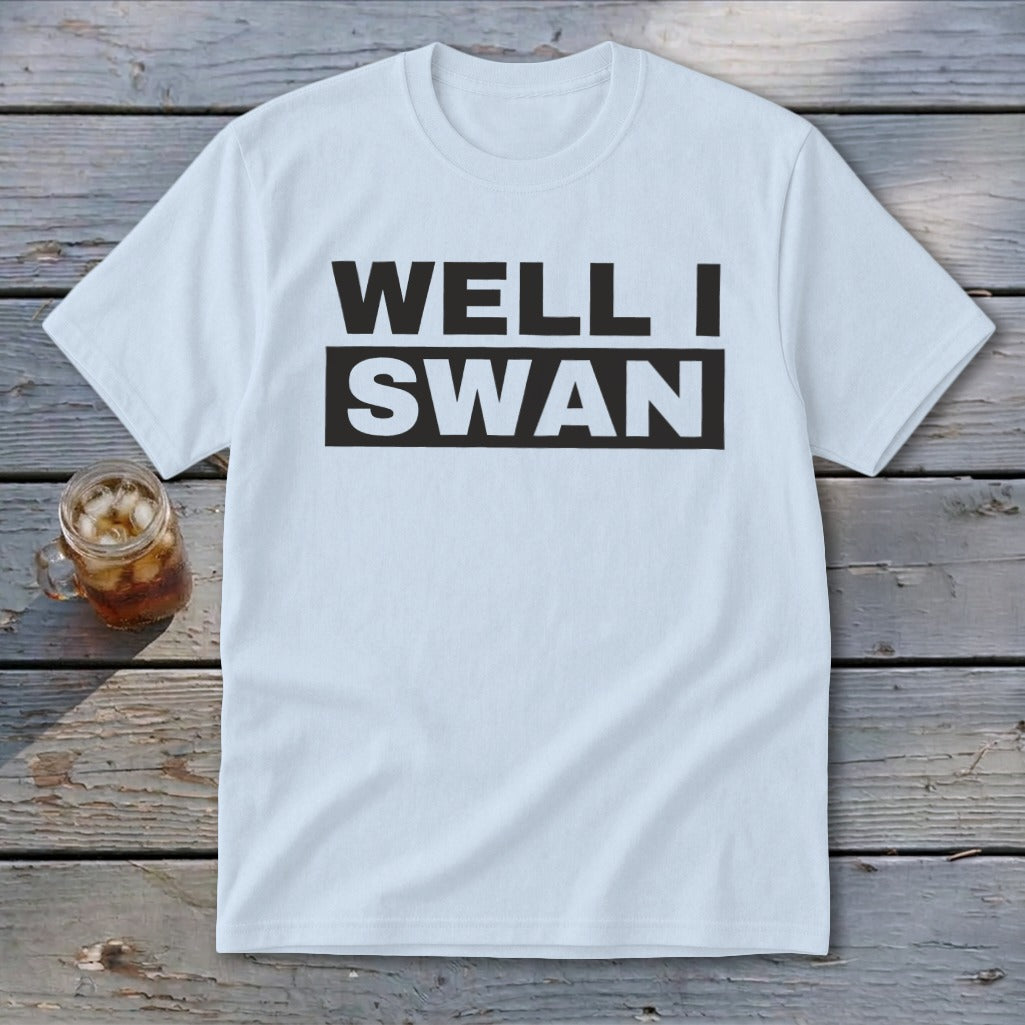 A white t-shirt featuring the black text WELL I SWAN lies on a wooden surface beside an iced beverage.