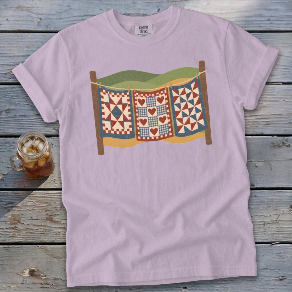 A light purple t-shirt featuring a retro illustration of three quilts hanging on a clothesline.