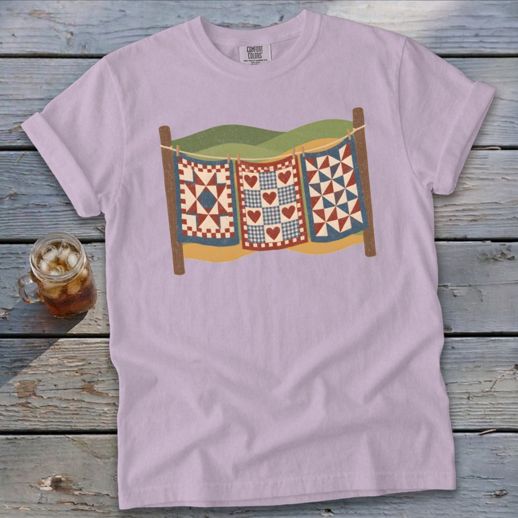 A light purple t-shirt featuring a retro illustration of three quilts hanging on a clothesline.
