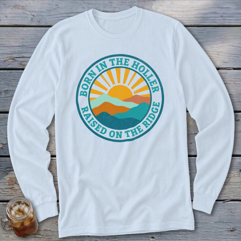 A white long-sleeved t-shirt features a circular graphic with mountains, a rising sun, and the text Born in the Holler, Raised on the Ridge.