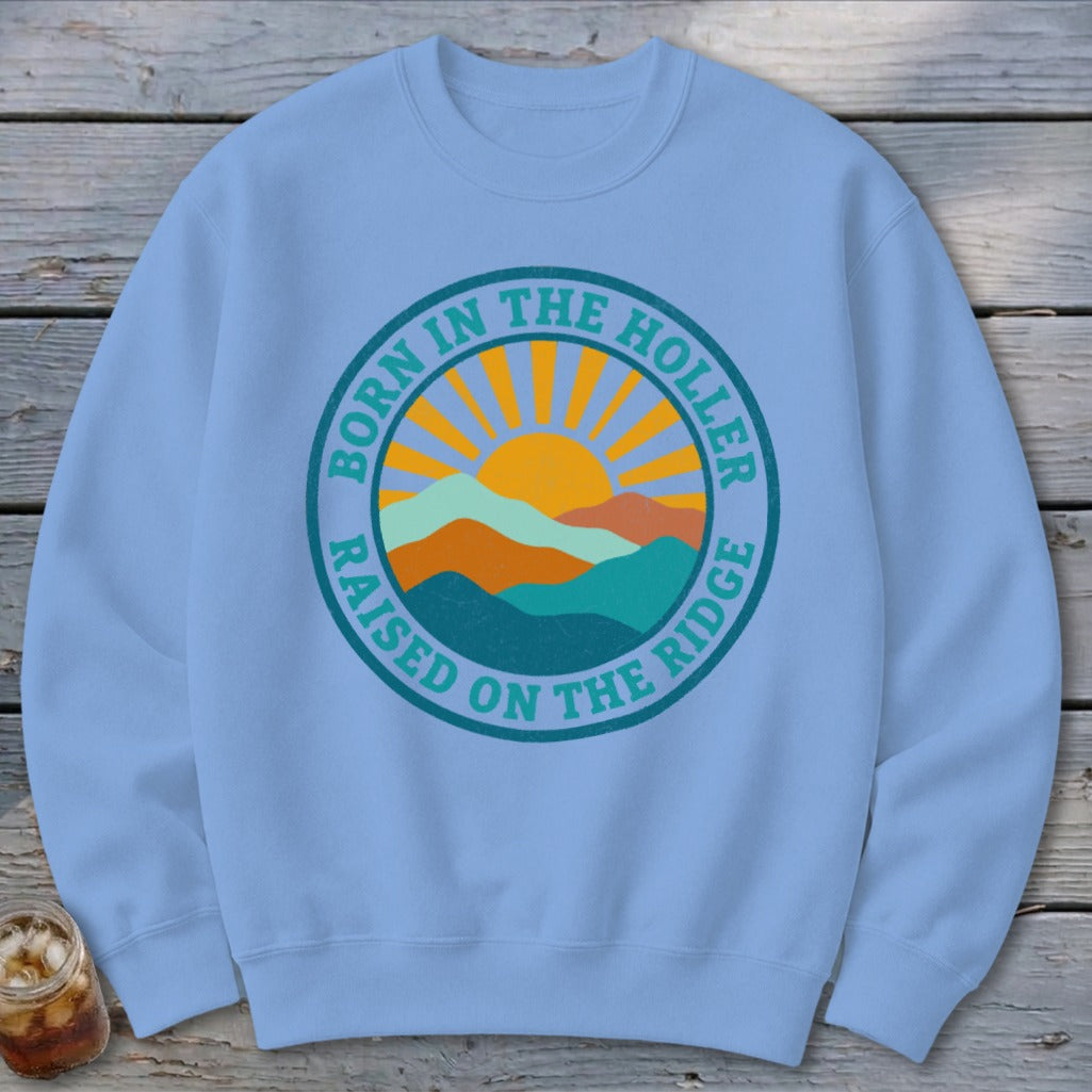 Light blue crewneck sweatshirt featuring a circular graphic with mountains, a rising sun, and text that reads BORN IN THE HOLLER RAISED ON THE RIDGE.