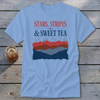 Light blue t-shirt featuring a graphic with the text Stars, Stripes & Sweet Tea laid over stylized red, white, and blue mountains.