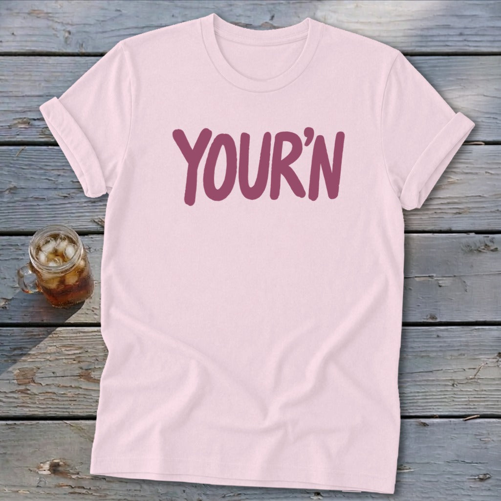 Light pink t-shirt with "YOUR'N" text in purple on a wooden surface.