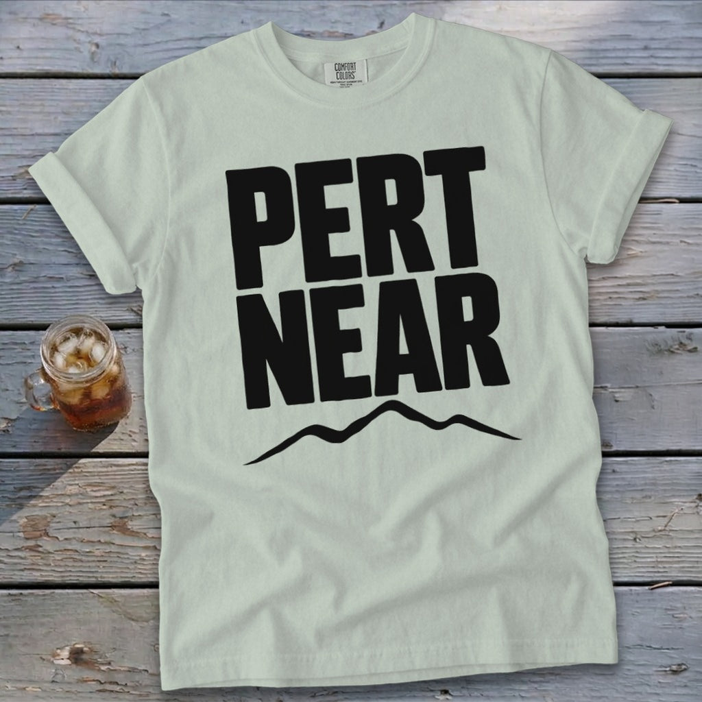 Light green t-shirt with black Pert Near text and mountain graphic lying on wooden surface.