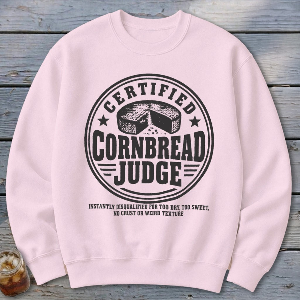A light pink crewneck sweatshirt featuring a black graphic with text reading Certified Cornbread Judge surrounding a cornbread illustration.