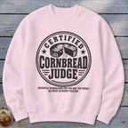 A light pink crewneck sweatshirt featuring a black graphic with text reading Certified Cornbread Judge surrounding a cornbread illustration.