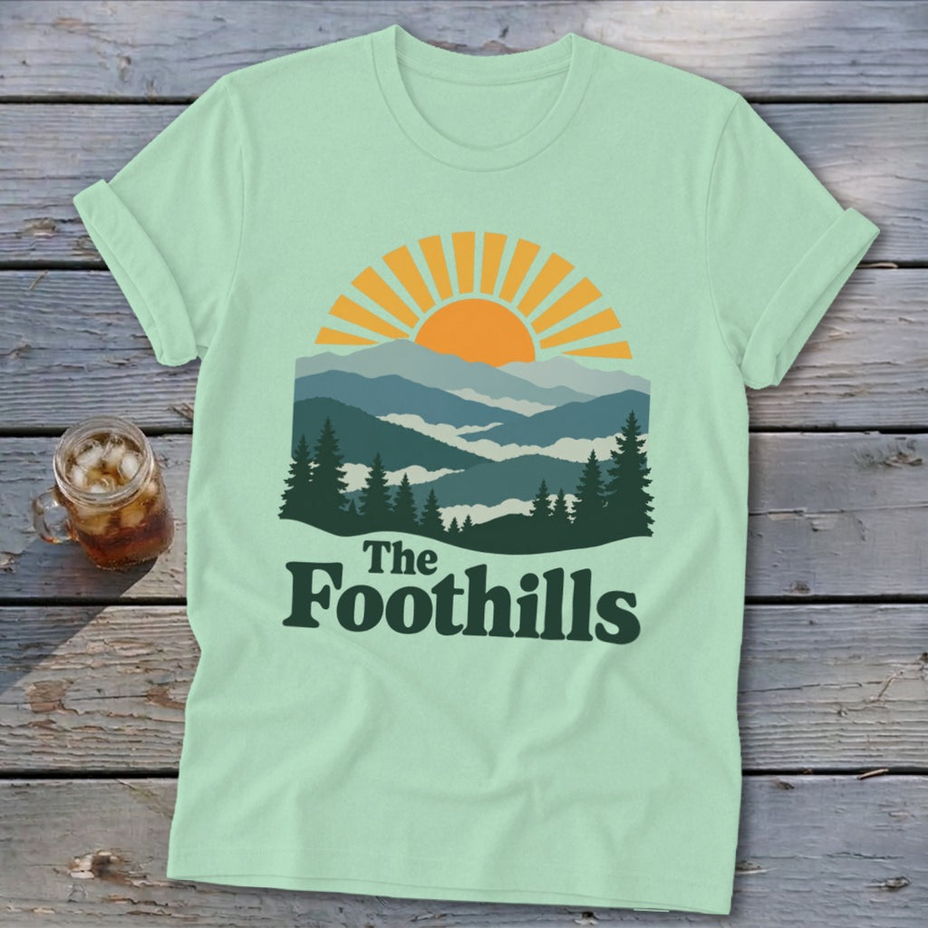 Mint t-shirt with sunrise over mountains graphic and "The Foothills" text.