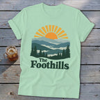 Mint t-shirt with sunrise over mountains graphic and "The Foothills" text.