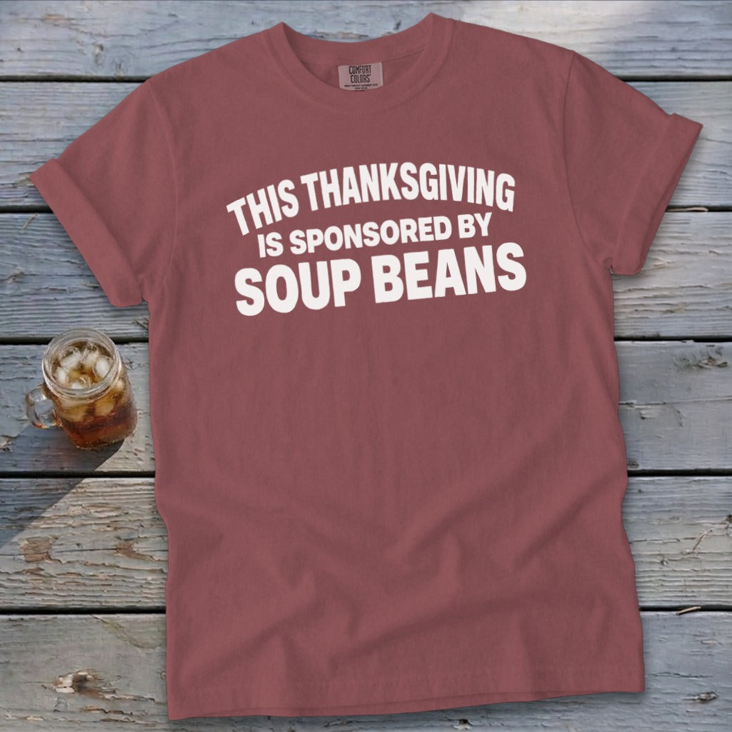 A mauve t-shirt with white text reading THIS THANKSGIVING IS SPONSORED BY SOUP BEANS laid on a wooden surface.
