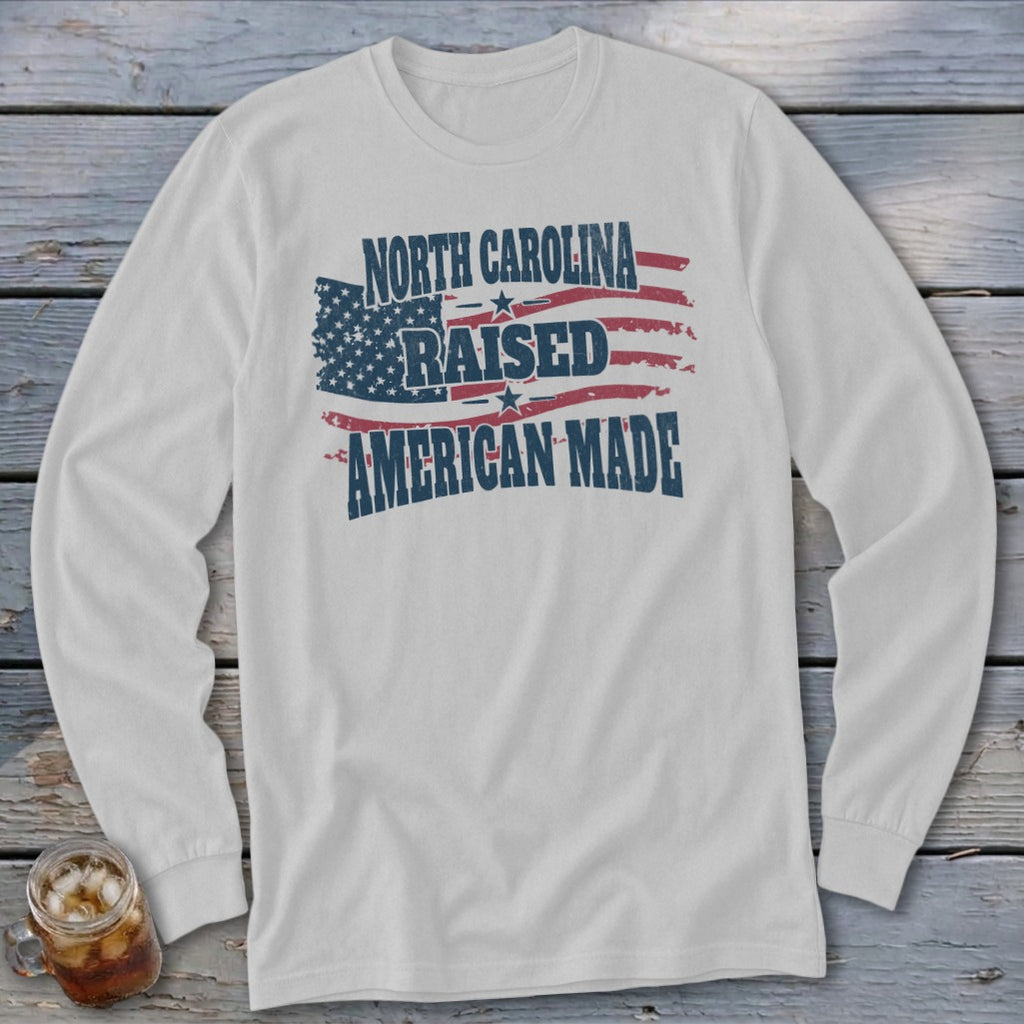 A light gray long-sleeve t-shirt featuring a red, white, and blue American flag graphic with the text NORTH CAROLINA RAISED AMERICAN MADE.