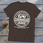 Brown short-sleeve t-shirt featuring a white graphic emblem reading Certified Cornbread Judge and a small drink beside it.