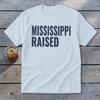 White t-shirt with "MISSISSIPPI RAISED" in dark blue text.