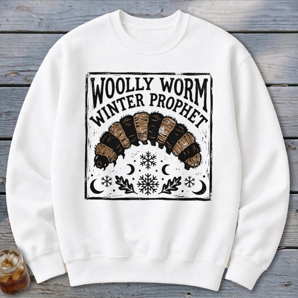A white crewneck sweatshirt features a black printed graphic of a woolly worm with winter motifs and text reading Woolly Worm Winter Prophet.