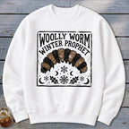 A white crewneck sweatshirt features a black printed graphic of a woolly worm with winter motifs and text reading Woolly Worm Winter Prophet.