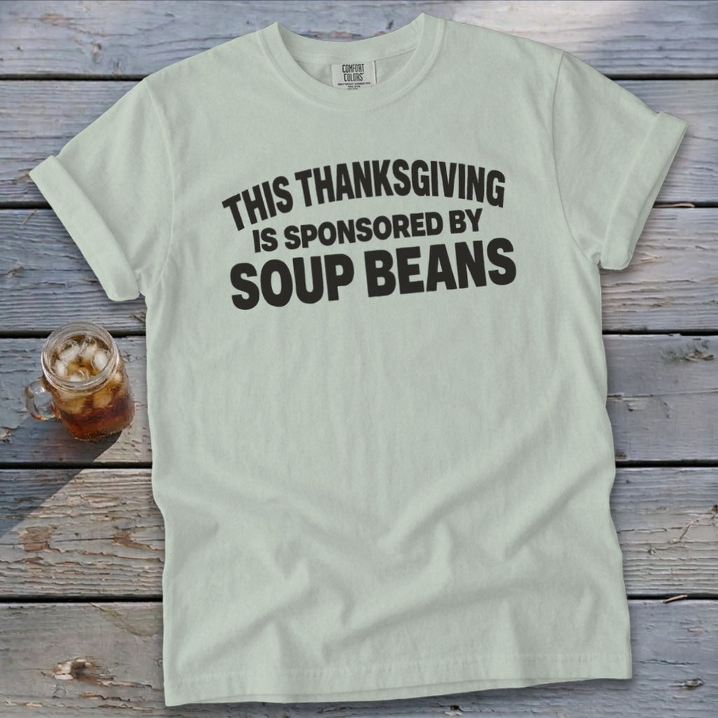 A light green t-shirt with black text reading THIS THANKSGIVING IS SPONSORED BY SOUP BEANS rests on a wooden surface next to a glass of iced tea.