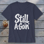 A dark blue V-neck t-shirt with white text reading Still A-Goin' on a wooden surface.