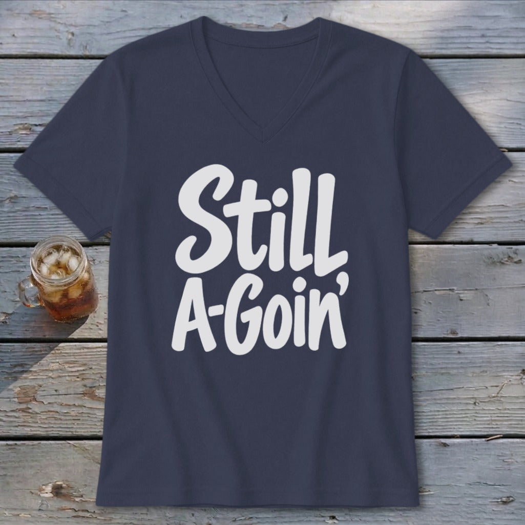 A dark blue V-neck t-shirt with white text reading Still A-Goin' on a wooden surface.