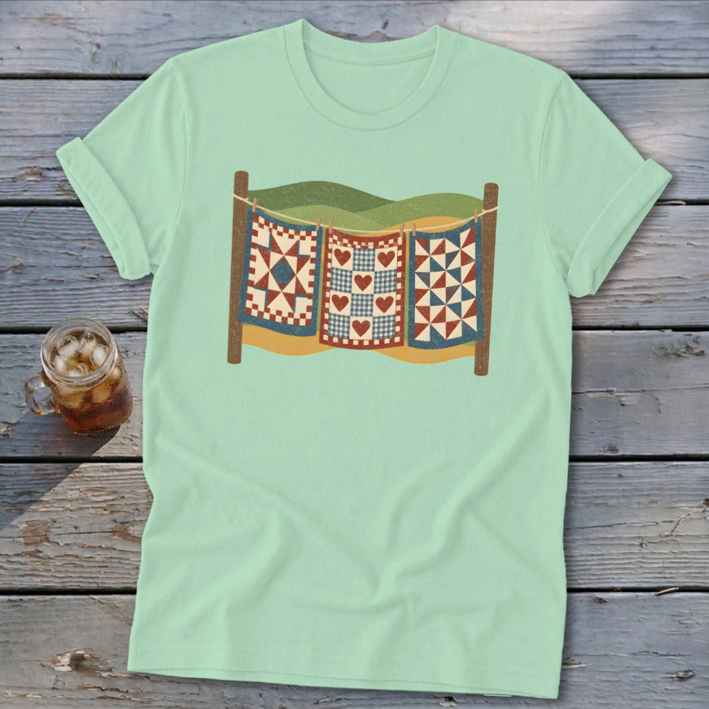 Light green t-shirt featuring a graphic of three patchwork quilts hanging on a clothesline over rolling hills.