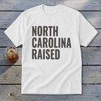 White t-shirt with bold gray text that reads "NORTH CAROLINA RAISED".