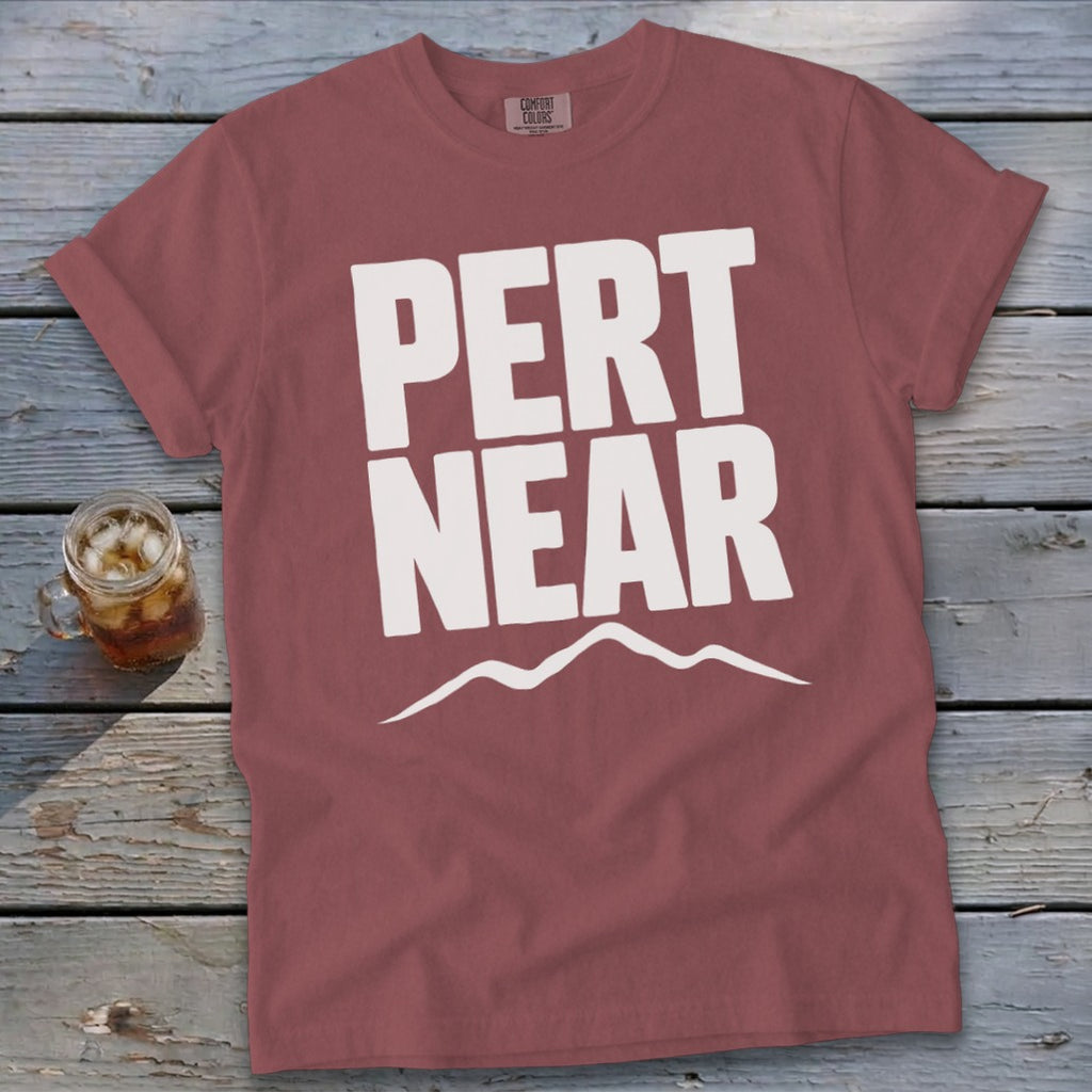 A dusty red t-shirt with white text reading PERT NEAR above a mountain range graphic, laid on a wooden background with a glass of iced tea.