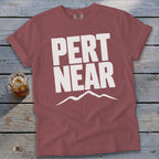 A dusty red t-shirt with white text reading PERT NEAR above a mountain range graphic, laid on a wooden background with a glass of iced tea.