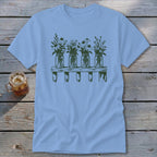 A light blue crewneck t-shirt featuring a green graphic of four mason jars filled with wildflowers on a wooden shelf, laid flat on a rustic wooden background next to a glass of iced tea.