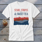 A white short-sleeve t-shirt features red and blue graphic text reading Stars, Stripes, & Sweet Tea above layered patriotic mountain silhouettes.