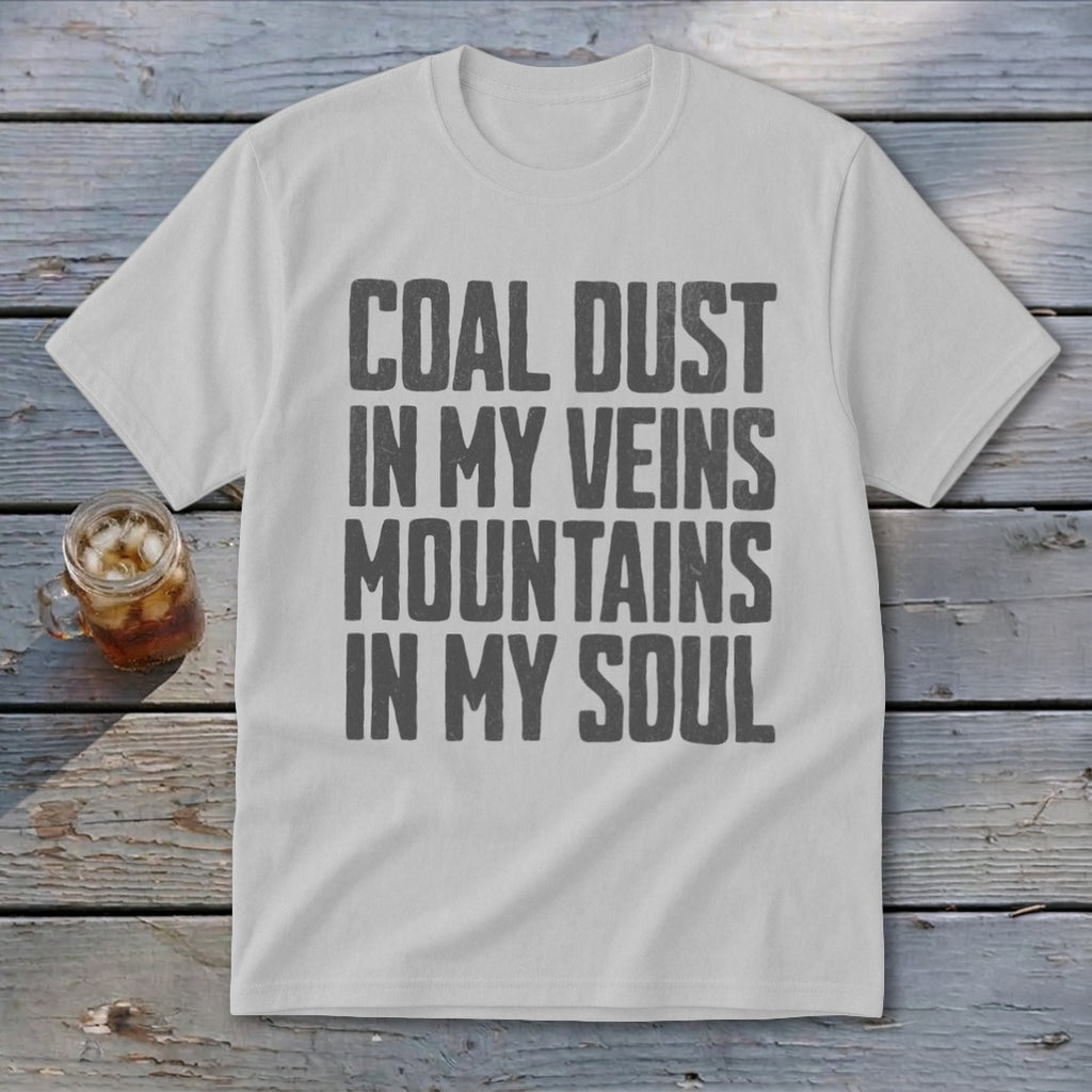 Light gray t-shirt with black block lettering reading COAL DUST IN MY VEINS MOUNTAINS IN MY SOUL, laid flat on a rustic wooden background next to a glass of iced tea.