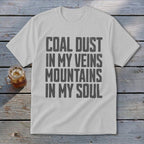 Light gray t-shirt with black block lettering reading COAL DUST IN MY VEINS MOUNTAINS IN MY SOUL, laid flat on a rustic wooden background next to a glass of iced tea.